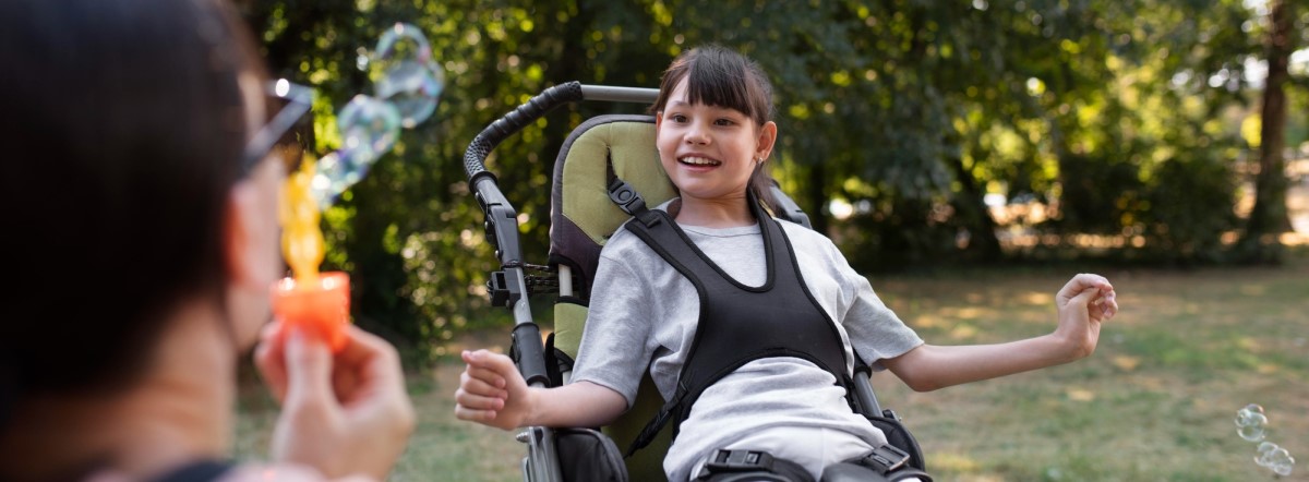 A happy girl in a wheelchair watches a person blow soap bubbles, radiating joy and positivity.